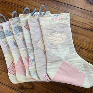 May include: Five quilted Christmas stockings arranged in a row, each featuring a light-colored fabric base with patchwork designs including floral patterns, red gingham, and a central tulip motif. Each stocking has a blue fabric hanging loop.