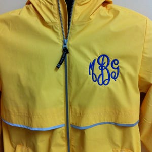 May include: A bright yellow hooded windbreaker jacket featuring a full-length grey front zipper with a black pull tab labeled Charles River. The jacket displays a blue embroidered monogram on the left chest and a horizontal grey reflective stripe.