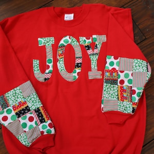 May include: A red sweatshirt with the word "JOY" appliqued on the front in patchwork fabric. The sleeves also feature patchwork fabric cuffs with red, green, and white patterns. The sweatshirt is a festive holiday garment.