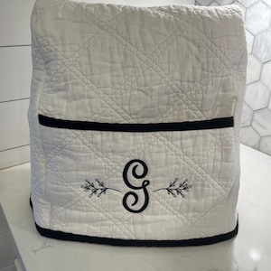 May include: A white quilted appliance cover featuring a black trim along the top pocket and bottom edge. The front is embroidered with a large black letter G centered between two delicate black botanical sprigs with small leaves.