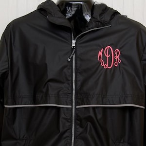 May include: A black hooded windbreaker jacket featuring a full-length silver zipper and a horizontal reflective grey stripe across the chest. The left chest area displays an embroidered monogram in bright pink cursive letters reading MDR.