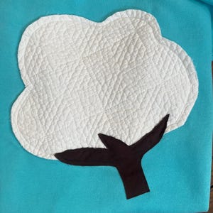May include: A turquoise sweatshirt featuring a white, quilted cotton plant design. The cotton plant has a fluffy, cloud-like appearance with a brown stem. The design is centered on the front of the sweatshirt.