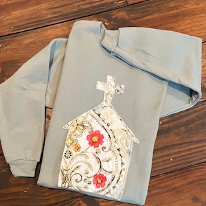 May include: A sage green sweatshirt with a church applique. The applique features a cross and a church building with a floral pattern in white, gold, and red. The sweatshirt is made of a soft material.
