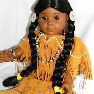 May include: A Native American doll with long black braids, wearing a brown suede dress with fringe and beaded jewelry. The doll has brown eyes and dark skin.