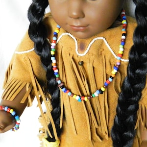 May include: A doll wearing a brown suede fringe outfit with a colorful beaded necklace and bracelet. The doll has long black braided hair.