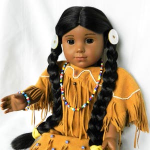 May include: A Native American doll with long black braids, wearing a brown suede dress with fringe and beaded jewelry. The doll has brown eyes and a neutral expression.