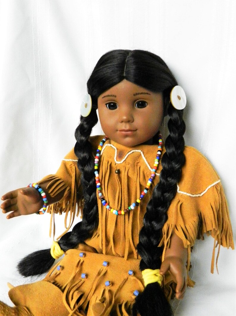 May include: A Native American doll with long black braids, wearing a brown suede dress with fringe and beaded jewelry. The doll has brown eyes and a neutral expression.