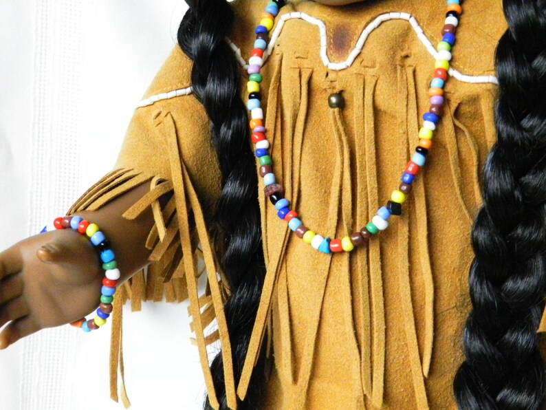 May include: A doll wearing a brown suede fringed outfit with a colorful beaded necklace and bracelet.