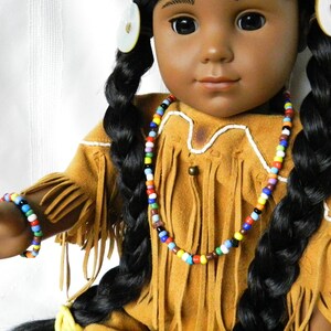 May include: A doll wearing a brown suede fringe jacket with white trim and a colorful beaded necklace and bracelet.