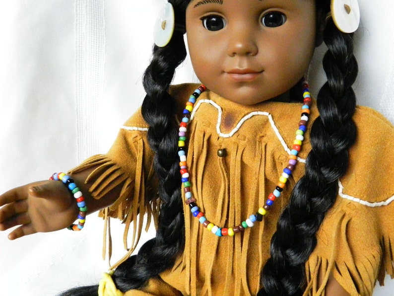 May include: A doll wearing a brown suede fringe jacket with white trim and a colorful beaded necklace and bracelet.