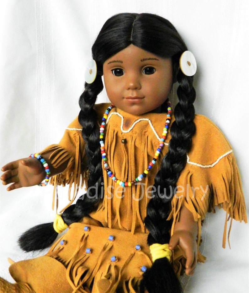 May include: A dark-skinned doll with long black braids wearing a brown suede dress with fringe and beaded jewelry. The doll has a white beaded headband and white earrings.
