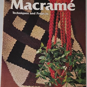 May include: A softcover book titled Macramé Techniques and Projects, A Sunset Book. The cover features a red macramé plant hanger holding a green plant, set against a background of a black and beige woven macramé wall hanging pattern.