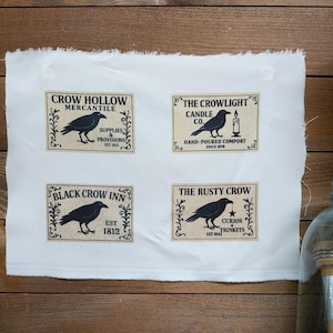 May include: A white fabric sheet featuring four rectangular labels with black crow illustrations and text. The labels read Crow Hollow Mercantile, The Crowlight Candle Co., Black Crow Inn, and The Rusty Crow, each with decorative borders.