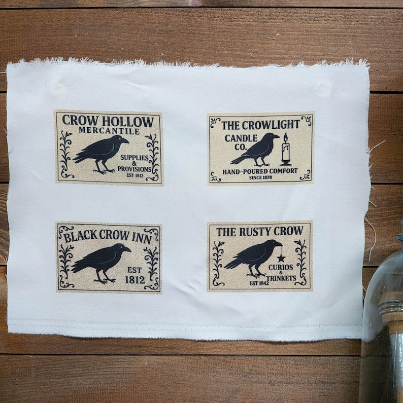 May include: A white fabric sheet featuring four rectangular labels with black crow illustrations and text. The labels read Crow Hollow Mercantile, The Crowlight Candle Co., Black Crow Inn, and The Rusty Crow, each with decorative borders.