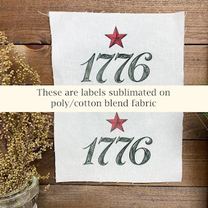 May include: Two white fabric labels with the number "1776" in dark green script, topped with a red star. The text "These are labels sublimated on poly/cotton blend fabric" is printed below the labels. The background is a wooden surface.