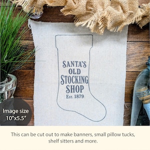 May include: A rectangular off-white fabric panel featuring a dark gray outline of a Christmas stocking with the text SANTA'S OLD STOCKING SHOP EST. 1879 printed in the center. The panel measures 10 inches by 5.5 inches and has raw, frayed edges.