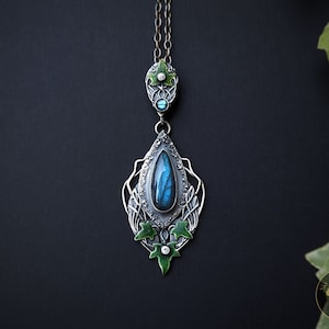 May include: A silver necklace with a teardrop-shaped pendant featuring a blue labradorite gemstone surrounded by green enamel ivy leaves. The pendant is attached to a silver chain with a smaller teardrop-shaped pendant with a blue gemstone and green enamel leaves.