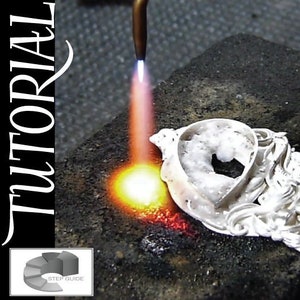 Könnte beinhalten: Nahaufnahme eines silbernen Anhängers, der mit einem Brenner gelötet wird. Der Anhänger hat ein detailliertes Design und liegt auf einer schwarzen Oberfläche. Der Text "Soldering - Jewelers Grade TUTORIAL" ist auf der linken Seite des Bildes sichtbar.