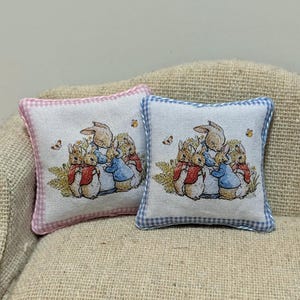 May include: Two miniature decorative pillows. Each pillow features a print of rabbits in a garden scene. One pillow has a pink gingham border, and the other has a blue gingham border. The pillows are on a tan surface.