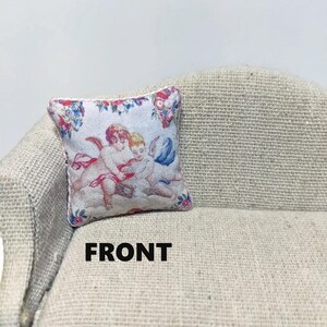 May include: A white pillow with a floral border and a print of two cherubs with wings. The pillow is on a light beige couch with the word "FRONT" printed below it.