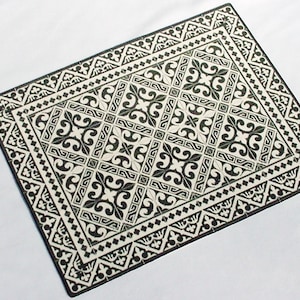 May include: A rectangular placemat featuring a high-contrast black and white pattern. The design consists of a central grid of repeating floral-inspired scrollwork motifs surrounded by a decorative border of geometric shapes and small diamond accents.