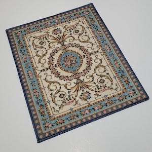 May include: A rectangular rug featuring a cream-colored center with a central blue medallion containing pink and green floral patterns. The border is light blue with repeating pink floral motifs and dark blue edges, showcasing intricate scrollwork.