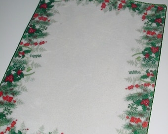 May include: Rectangular placemat with a white center and a border featuring green pine needles, red berries, and green leaves. The placemat has a green stitched edge, creating a festive holiday theme.