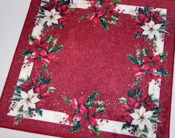 May include: A square, red Christmas-themed decorative cloth with a glittery texture. The cloth features a white border with poinsettias and holly arranged in each corner. The poinsettias are red and white, with green holly leaves.