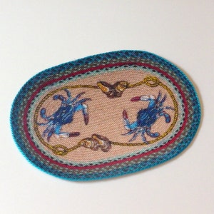 May include: Oval-shaped woven rug with a blue and white crab design on a beige background. The rug has a blue, red, and green border.