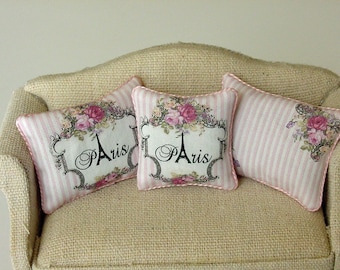 May include: Three decorative throw pillows with a pink and white striped pattern and a floral design featuring pink roses and the word "Paris" in a script font. The pillows are on a beige couch.