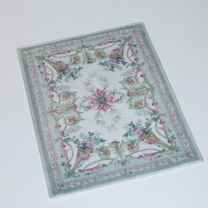 May include: A white rug with a green and pink floral pattern. The rug has a decorative border with a scalloped edge.