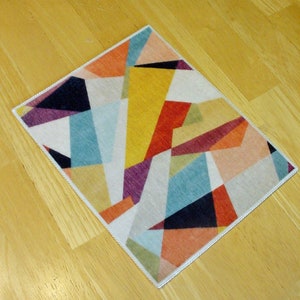 May include: A colorful geometric rug with a white background and a variety of colors including red, orange, yellow, green, blue, purple, and black. The rug has a white border.