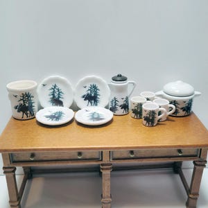 May include: A miniature white ceramic dining set with a moose and tree design. The set includes plates, cups, a pitcher, a pot with a lid, and a container. The moose and tree design is in dark blue.