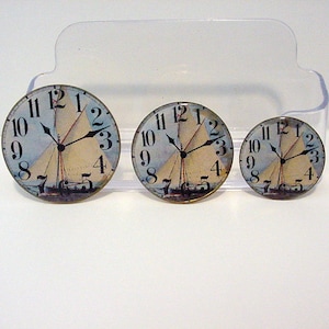 May include: Three round clock faces with a sailboat design. The clock faces are white with black numbers and hands. The largest clock face is 2.5 inches in diameter, the middle clock face is 2 inches in diameter, and the smallest clock face is 1.5 inches in diameter.
