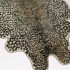 May include: A miniature rug with a leopard print design. The rug features a beige background with black spots and a brown rope-like border. The rug is shaped like an animal hide.