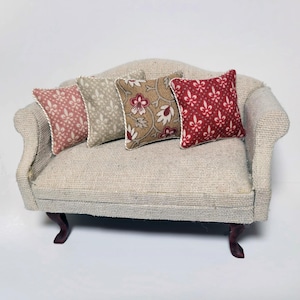 May include: Miniature beige sofa with four patterned pillows. The pillows feature floral and geometric designs in shades of red, tan, and beige. The sofa has dark brown legs and a textured fabric.