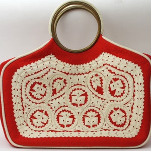 Puede incluir: Bolso de mano de fieltro rojo con un dise&ntilde;o bordado blanco y un asa grande redonda de lat&oacute;n.