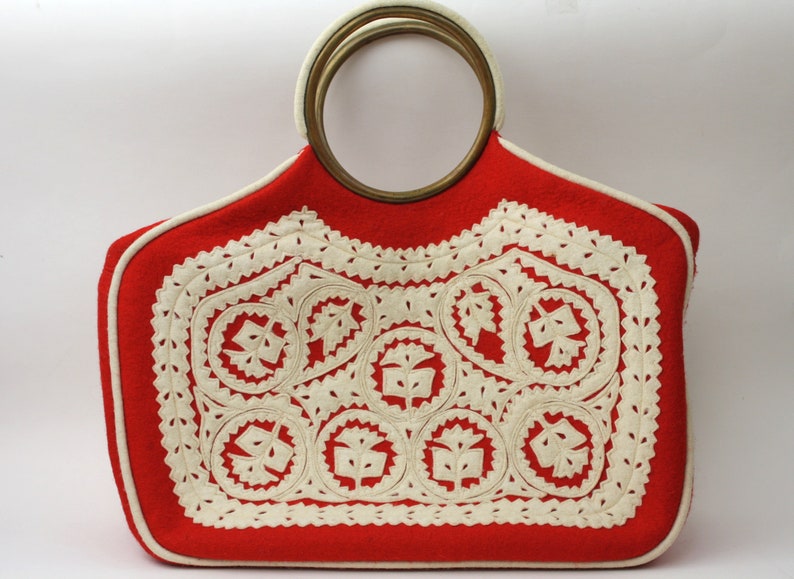 Puede incluir: Bolso de mano de fieltro rojo con un dise&ntilde;o bordado blanco y un asa grande redonda de lat&oacute;n.