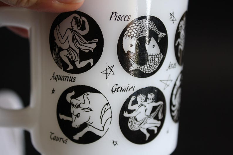 May include: White ceramic mug featuring black zodiac illustrations. The mug displays various zodiac signs like Aquarius, Pisces, Taurus, and Gemini. Each sign is within a black circle, with the sign's name below it.