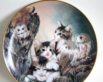 May include: A decorative porcelain plate featuring a whimsical scene with four cats and two owls. The cats are depicted in various poses, with one cat perched on a tree branch, another cat looking up at the viewer, and two cats cuddling together. The owls are perched on branches, with one owl looking at the viewer and the other owl looking down at the cats. The plate has a gold rim and a white background.