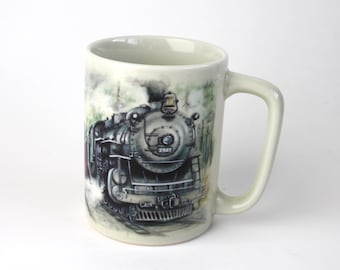 May include: A white ceramic mug with a black and white image of a steam locomotive train with the number 2947 on the side. The train is surrounded by a green and white watercolor background.