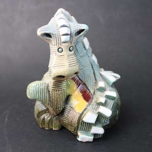 May include: A ceramic dragon figurine in shades of green, tan, and white. The dragon has a textured, patterned design and small blue eyes. A patch of red, yellow, and brown adds a pop of colour. The figurine is a decorative item.