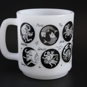 May include: White ceramic mug featuring black zodiac symbols, including Pisces, Aries, and Taurus. The mug has a rounded shape with a handle and is decorated with star accents. A vintage-style design.