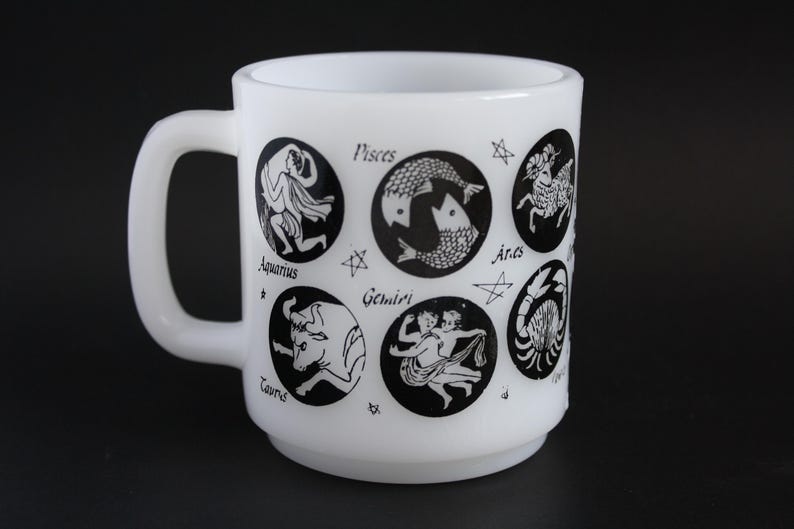 May include: White ceramic mug featuring black zodiac symbols, including Pisces, Aries, and Taurus. The mug has a rounded shape with a handle and is decorated with star accents. A vintage-style design.