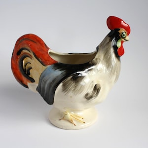 Peut inclure: Un pot à lait en céramique en forme de coq, doté d'un corps blanc cassé brillant avec des coups de pinceau noirs peints à la main, une crête et des plumes de queue rouge vif, et un bec jaune. Le récipient repose sur une base circulaire.