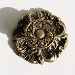 Peut inclure: Un bouton décoratif en métal doré avec un motif floral. Le bouton a un centre surélevé avec un motif circulaire.