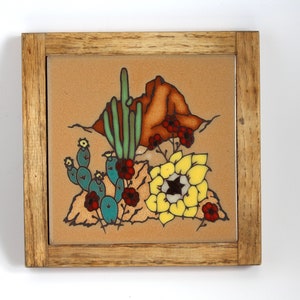 May include: A framed ceramic tile with a desert scene featuring a cactus, a yellow flower, and a red rock formation. The tile is surrounded by a wooden frame.
