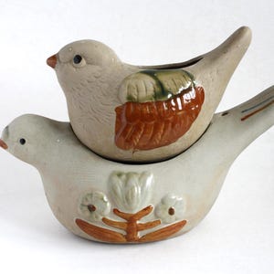May include: Two ceramic bird ornaments, one stacked on the other. The top bird is beige with brown and green wing details. The bottom bird is light grey with floral and brown accents. The birds have small black eyes and orange beaks.