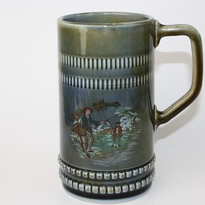 May include: A green and blue ceramic mug with a white rim and handle. The mug features a scene of two men fishing in a river with trees and a cloudy sky in the background.