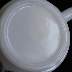 May include: A white ceramic mug with a rounded base and a small handle. The bottom of the mug has a slightly recessed center and a raised rim. The number "26" is imprinted on the bottom.